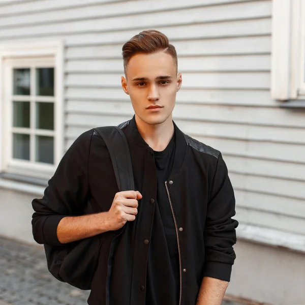 Joven guapo con peinado en una chaqueta con una mochila cerca de una casa de madera . — Foto de Stock