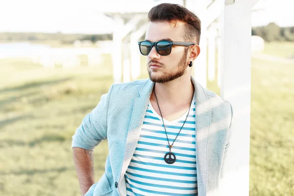Handsome hipster man in sunglasses with a beard in a trendy suit and shirt on the beach. — Stock Photo, Image