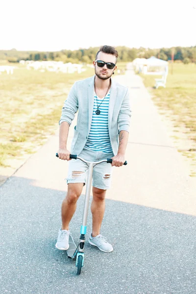 Beau jeune homme hipster dans une veste élégante en short déchiré chevauchant un scooter . — Photo
