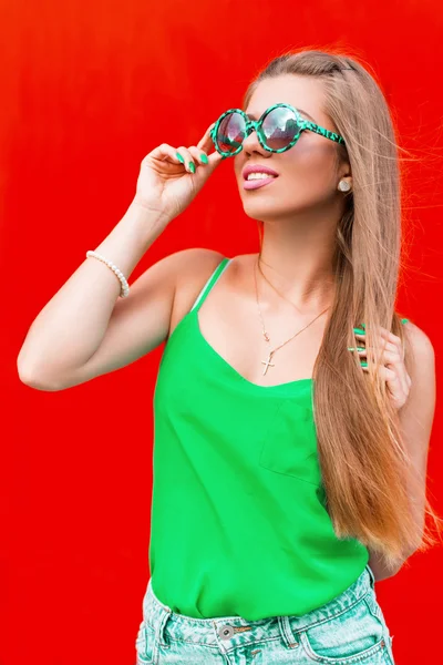 Portrait lumineux d'été de jeune belle femme gaie avec un sourire aux lèvres roses dans des lunettes de soleil rondes vertes près du mur rouge — Photo