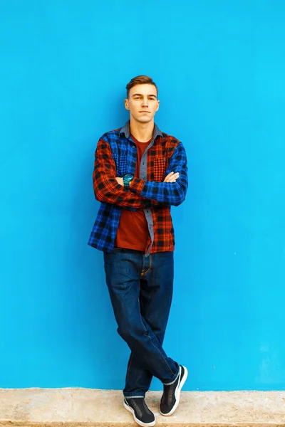 Stijlvolle knappe man in een modieuze shirt, sneakers en jeans staat in de buurt van een heldere blauwe muur. — Stockfoto