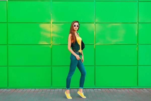 Elegante mujer hermosa en una camiseta amarilla, jeans y zapatillas de deporte posando cerca de la pared verde . —  Fotos de Stock