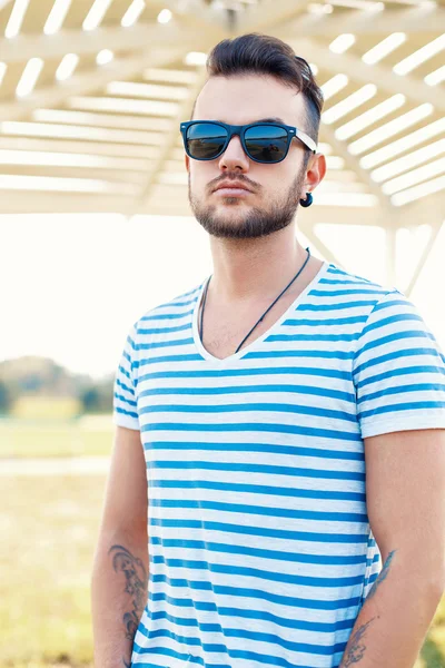 Mladý pohledný hipster chlap s vousy ve stylovém tričku a slunečními brýlemi ve slunečném letním dni na pláži. — Stock fotografie