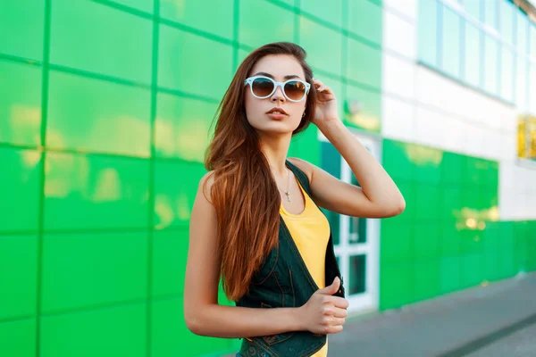 Hermosa mujer joven en ropa elegante brillante se encuentra cerca de un edificio moderno . — Foto de Stock