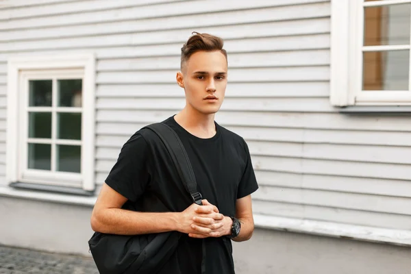 Guapo chico con estilo en una camiseta negra con una mochila de pie cerca de una casa de madera . — Foto de Stock
