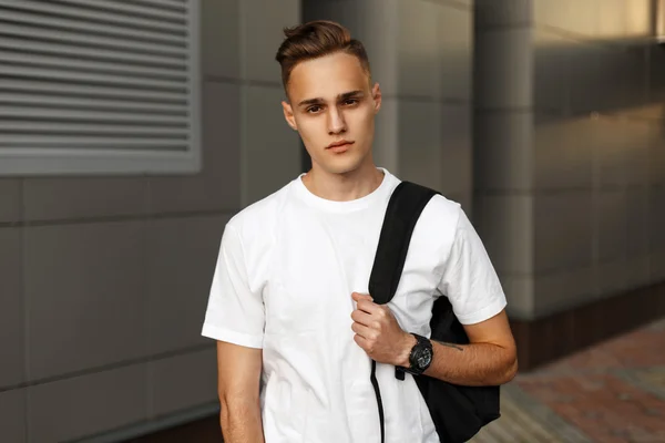 Beau hipster homme en T-shirt blanc avec un sac à dos sur le fond d'un bâtiment moderne . — Photo