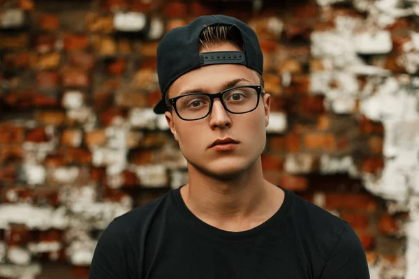 Tipo guapo con gafas en una gorra de béisbol negro y camiseta negra de pie sobre un fondo de pared de ladrillo — Foto de Stock