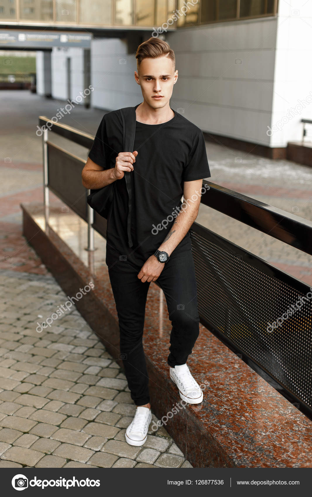 Handsome hipster man in stylish black t-shirt, pants and white sneakers  with a backpack standing near the modern buildin Stock Photo by ©alonesdj  126877536