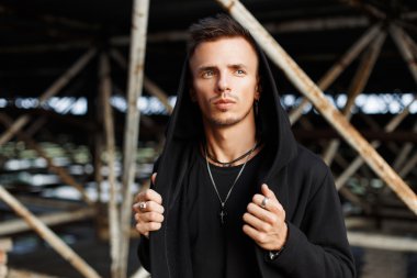 Metal konstrüksiyon bir arka plan karşı poz bir kukuleta ile siyah moda giysiler içinde şık yakışıklı