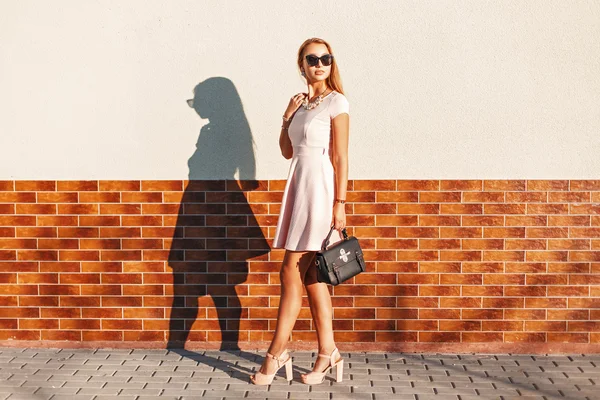 Stylisches junges Mädchen mit Sonnenbrille in rosa Rock mit schwarzer Tasche in der Nähe der Ziegelwand. — Stockfoto