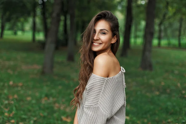 Χαρούμενο όμορφο κορίτσι με ένα όμορφο χαμόγελο σε ένα γκρίζο πουλόβερ κοντά στα δέντρα στο πάρκο — Φωτογραφία Αρχείου