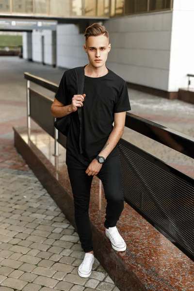 Hombre hipster guapo en elegante camiseta negra, pantalones y zapatillas blancas con una mochila de pie cerca del edificio moderno — Foto de Stock