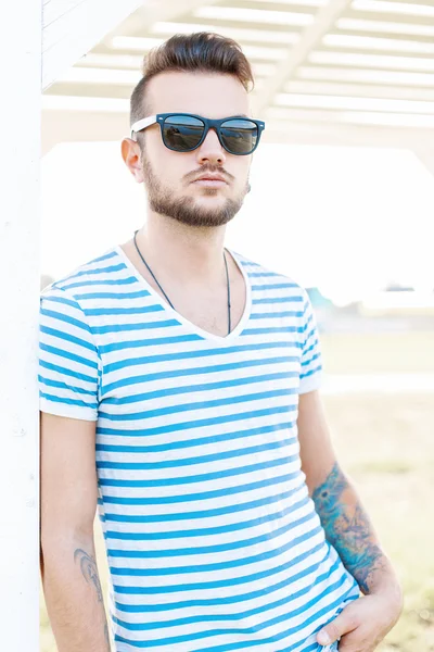 Handsome hipster man with a beard in sunglasses standing on the beach on a sunny day. — Stock Photo, Image