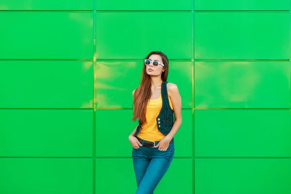 Hermosa mujer en una camiseta amarilla y chaqueta de mezclilla de pie cerca de una pared de metal verde . —  Fotos de Stock