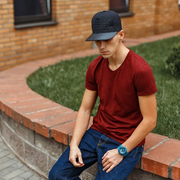 Trendy knappe jongen in een zwarte Baseballcap zit in de buurt van een baksteen gebouw. — Stockfoto