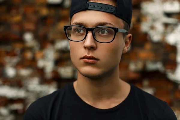 Joven hombre con estilo con gafas y una gorra negra se encuentra cerca de una pared de ladrillo rojo — Foto de Stock