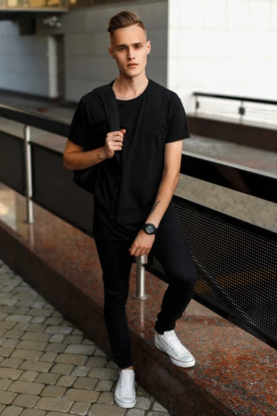 Stilig ung man i snygga svarta kläder och vita skor med en ryggsäck som poserar nära ett räcke — Stockfoto