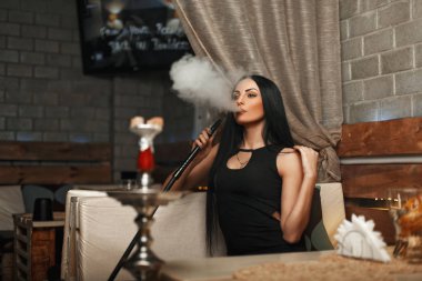 The beautiful girl smokes a hookah and lets the smoke. Woman resting in a bar
