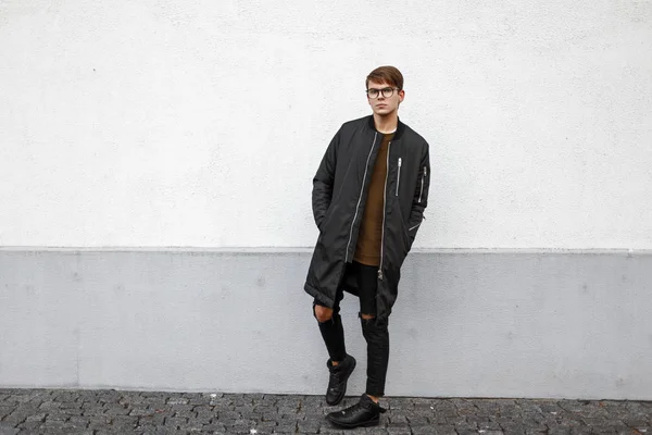 Young model guy with glasses in a black jacket and torn jeans stands near the wall Royalty Free Stock Images