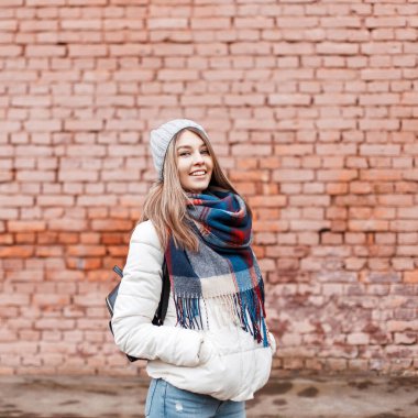 Neşeli güzel genç kız beyaz bir ceket ve tuğla duvarın yanında siyah bir çanta ile renkli bir eşarp tatlı bir gülümseme