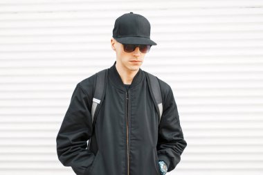 Beysbol şapkası ve beyaz metal duvarının yakınında bir sırt çantası ile siyah ceketli genç yakışıklı çocuk