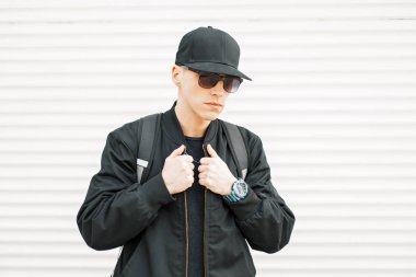 Amerikan yakışıklı genç bir siyah beyzbol şapkası ve siyah güneş gözlüğü ve beyaz metal duvarının yakınında şık bir ceket