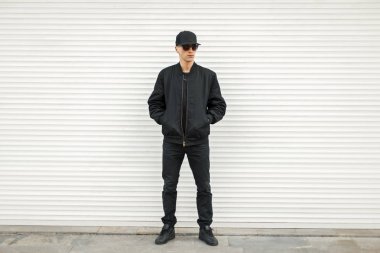 Siyah modern ceket, beyzbol şapkası ve siyah spor ayakkabı beyaz perdeleri yakınındaki gözlüklü yakışıklı adam