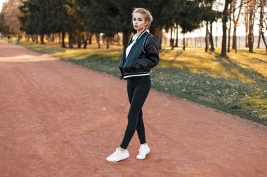 Güzel genç kadın şık bir ceket, siyah kot ve beyaz ayakkabı parkta yürüyüş.