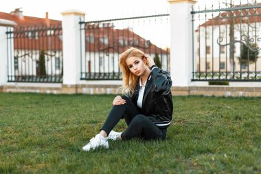 Genç güzel kız öğrenci moda ceket, siyah kot ve beyaz spor ayakkabı çimenlerin üzerinde oturur