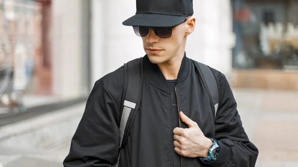 Jovem na moda óculos de sol em um boné preto e casaco com um saco na cidade — Fotografia de Stock