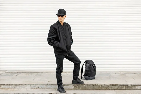 Przystojny stylowy mężczyzna w czarną kurtkę i trampki stoi w pobliżu plecak — Zdjęcie stockowe