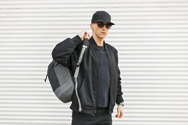 Jovem elegante em roupas modernas pretas com uma mochila perto de uma parede branca — Fotografia de Stock