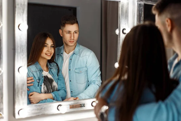 Hermosa joven pareja alegre sonriendo en jeans ropa mirando en el espejo con luces — Foto de Stock