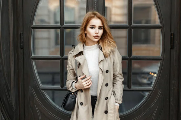 Красивая модная молодая блондинка в пальто с сумкой возле двери отеля — стоковое фото