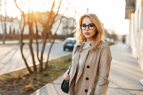Beautiful young woman in stylish glasses and trendy coat with handbag in the day of spring at sunset