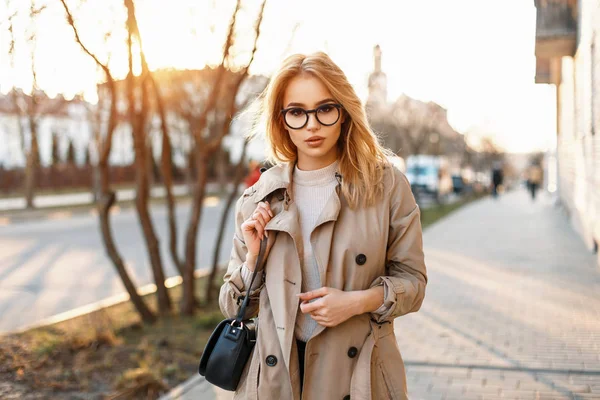 Belle jeune fille hipster avec des lunettes de soleil dans un manteau avec un sac à pied dans la ville de printemps au coucher du soleil — Photo