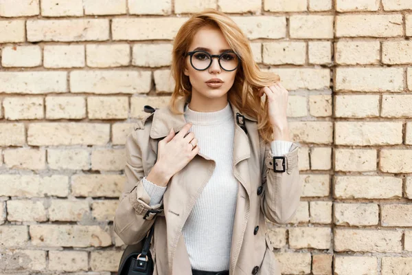 Mulher hipster jovem bonita com óculos de sol no casaco com bolsa preta perto da parede de tijolo — Fotografia de Stock