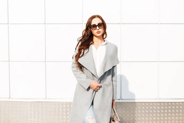 Jeune femme en lunettes de soleil tendance et manteau élégant avec sac près du mur blanc par une journée ensoleillée — Photo