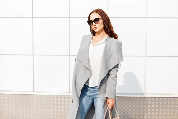 Élégante belle femme en lunettes de soleil en manteau gris avec sac près du mur blanc — Photo