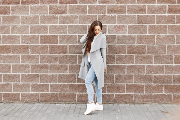 Schöne modische Mädchen in einem grauen Mantel und Jeans in der Nähe einer Ziegelwand — Stockfoto