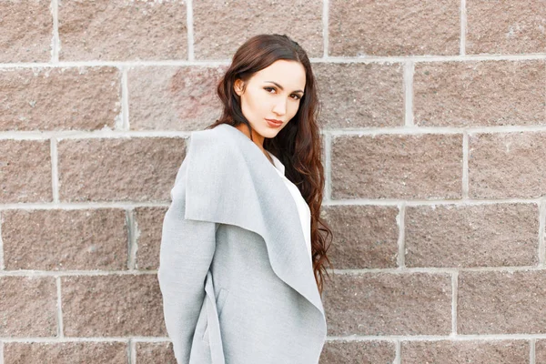 Hermosa chica en un abrigo gris de moda cerca de una pared de ladrillo —  Fotos de Stock