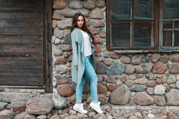 Elegante bella ragazza in un cappotto alla moda e scarpe da ginnastica bianche vicino al muro di pietra — Foto Stock