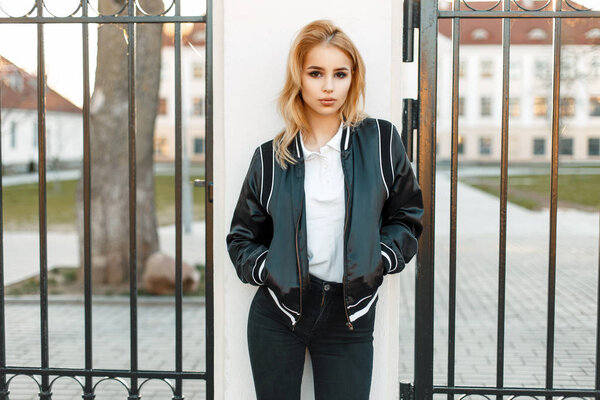 Молодая студентка в черной куртке в колледже возле забора
