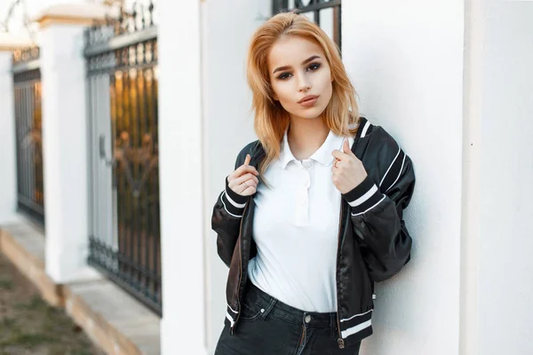 Mulher loira jovem bonita em jaqueta preta e camisa de pólo branco perto da parede branca — Fotografia de Stock