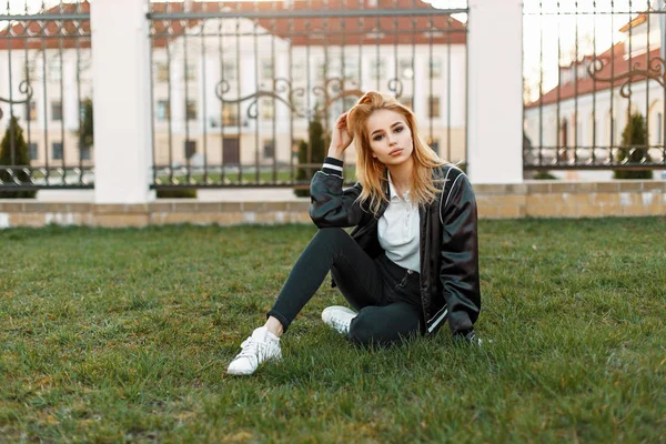 スタイリッシュなジャケットと白いスニーカーと黒のジーンズで美しい若い女子学生が草の上に座っています。 — ストック写真