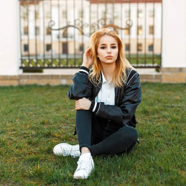 Mulher loira bonita em uma jaqueta preta e jeans preto com sapatos brancos descansando na grama — Fotografia de Stock