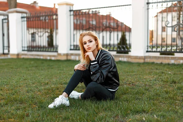 Beautiful young girl in a bomber jacket in black jeans and white shoes sitting on the grass.