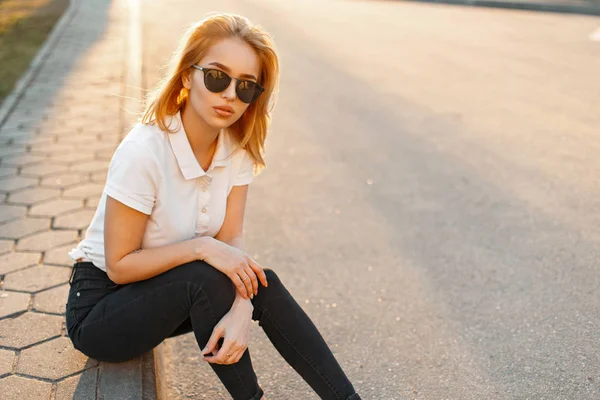 Молодая красивая женщина в белой рубашке для поло в солнечных очках сидит на обочине дороги на закате — стоковое фото