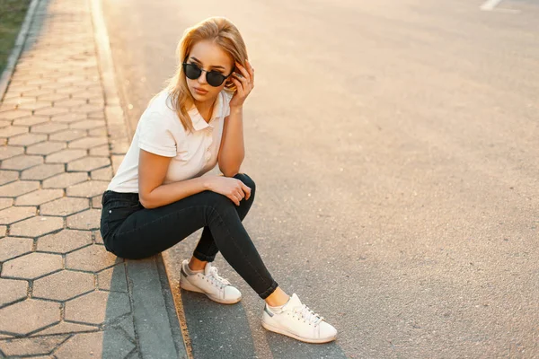 Красива жінка в модних сонцезахисних окулярах в білій сорочці поло та чорних джинсах з білим взуттям сидить на асфальті під час заходу сонця — стокове фото