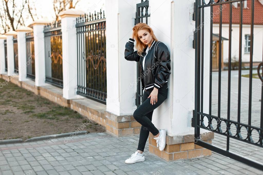 Beautiful young woman in a bomber jacket with black jeans and white shoes is standing near the fence of the collegiate